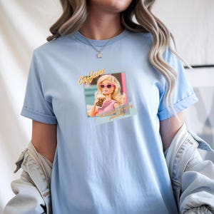 Puede incluir: Camiseta azul claro con un gr&aacute;fico de una mujer rubia con gafas de sol rosas sosteniendo una bebida. El texto "Caffeine" est&aacute; encima de la imagen, y "...is my personality" debajo. La camiseta tiene mangas cortas.