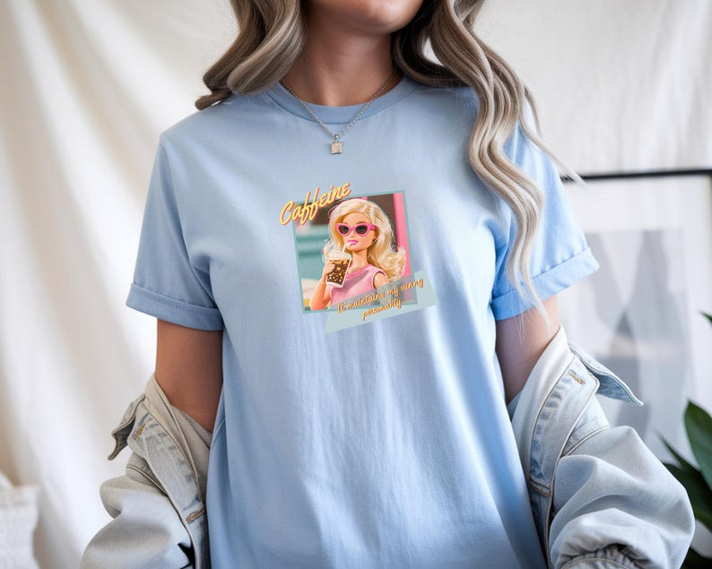 Puede incluir: Camiseta azul claro con un gr&aacute;fico de una mujer rubia con gafas de sol rosas sosteniendo una bebida. El texto "Caffeine" est&aacute; encima de la imagen, y "...is my personality" debajo. La camiseta tiene mangas cortas.