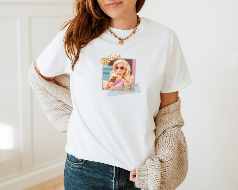 Puede incluir: Camiseta blanca con un gr&aacute;fico de una mujer rubia con gafas de sol rosas, sosteniendo una bebida. El texto "Caffeine" est&aacute; encima de la imagen, con "...is my personality" debajo. La camiseta la lleva una persona con vaqueros azules y una rebeca beige.