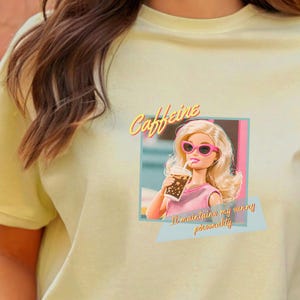 Puede incluir: Camiseta amarillo claro con un gr&aacute;fico de una mu&ntilde;eca con gafas de sol rosas y sosteniendo una bebida. Tambi&eacute;n se muestra el texto "Caffeine" y "It maintains my sunny personality".