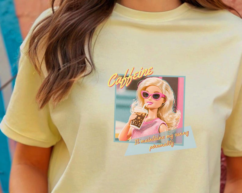 Puede incluir: Camiseta amarillo claro con un gr&aacute;fico de una mu&ntilde;eca con gafas de sol rosas y sosteniendo una bebida. Tambi&eacute;n se muestra el texto "Caffeine" y "It maintains my sunny personality".
