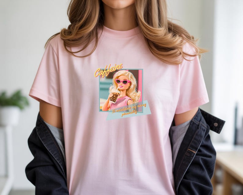 Puede incluir: Camiseta rosa claro con un estampado de una mu&ntilde;eca rubia con gafas de sol rosas y una bebida. El texto "Caffeine" est&aacute; encima de la mu&ntilde;eca, y "I'm maintaining my wing personality" est&aacute; debajo. Estilo casual.