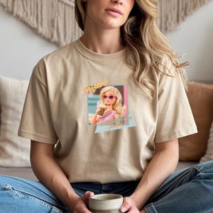 Puede incluir: Camiseta beige con un gr&aacute;fico retro de una mujer de dibujos animados con gafas de sol sosteniendo una bebida. El gr&aacute;fico incluye la palabra "Caffeine" y la frase "the ultimate pick me up". La camiseta es de material suave.