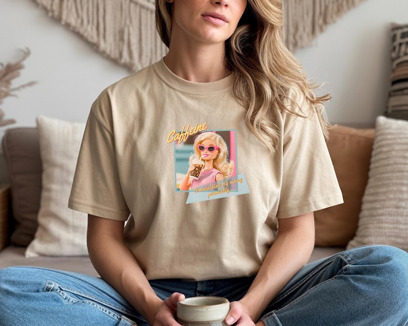 Puede incluir: Camiseta beige con un gr&aacute;fico retro de una mujer de dibujos animados con gafas de sol sosteniendo una bebida. El gr&aacute;fico incluye la palabra "Caffeine" y la frase "the ultimate pick me up". La camiseta es de material suave.