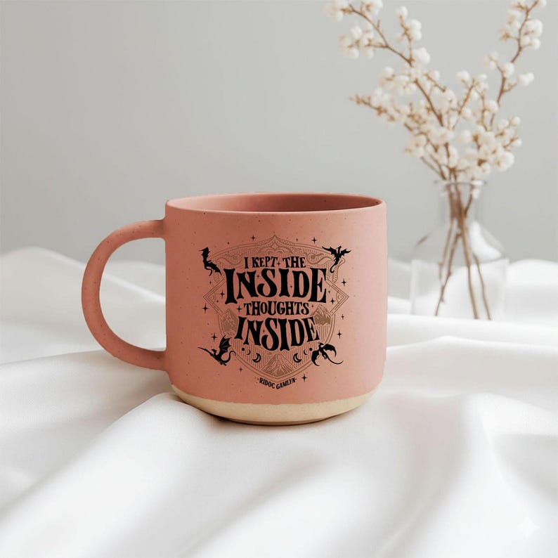 May include: A matte dusty pink ceramic mug with a speckled texture and a cream-colored base. The front features black text that reads I KEPT THE INSIDE THOUGHTS INSIDE, surrounded by a decorative shield, small stars, and dragon silhouettes.