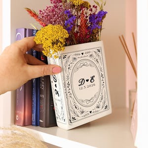 May include: A white ceramic book-shaped vase filled with colorful dried flowers. The vase has a black floral design and the initials "D & E" with the date "12.3.2026". The vase is on a white shelf next to a row of books.