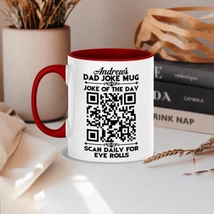 May include: A white ceramic mug with a red interior and handle. The front features black text reading Andrew's DAD JOKE MUG, JOKE OF THE DAY, and SCAN DAILY FOR EYE ROLLS, surrounding a central black and white QR code for scanning purposes.