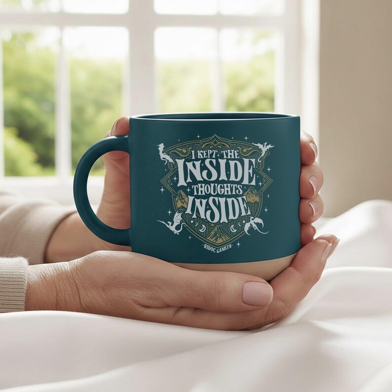 May include: A dark teal ceramic mug with a light-colored base. The front features a decorative gold and white emblem with the text "I KEPT THE INSIDE THOUGHTS INSIDE" and "RIDOC GAMLEN" surrounded by small dragon illustrations and star accents.