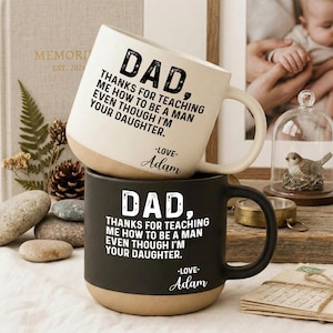 May include: Two ceramic mugs, one white and one black, stacked on top of each other. Each mug has a tan base and a large handle. The white mug reads "DAD, THANKS FOR TEACHING ME HOW TO BE A MAN EVEN THOUGH I'M YOUR DAUGHTER. -LOVE- Adam". The black mug has the same text.