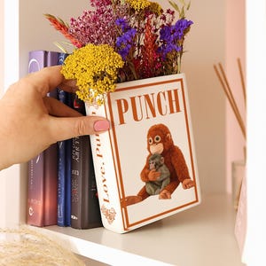 May include: A white ceramic book-shaped vase with the word "PUNCH" and an illustration of a monkey holding a baby monkey. The vase is filled with colorful dried flowers. The side of the vase reads "Love, Pur".