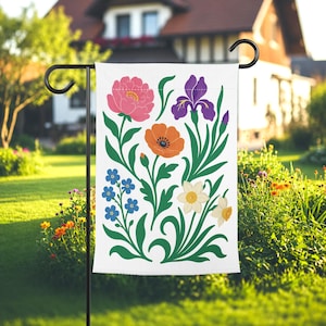 Op de afbeelding: Een witte tuinvaandel met een bloemenontwerp. Het heeft een roze pioenroos, paarse iris, oranje klaproos, blauwe vergeet-mij-nietjes en witte narcissen, tegen een achtergrond van groene bladeren en stelen.