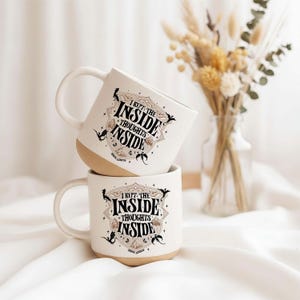 May include: Two off-white ceramic mugs stacked vertically, each featuring a black and gold graphic design with the text I KEPT THE INSIDE THOUGHTS INSIDE. The mugs have a smooth handle and a natural, unglazed tan base, showcasing a detailed illustration.