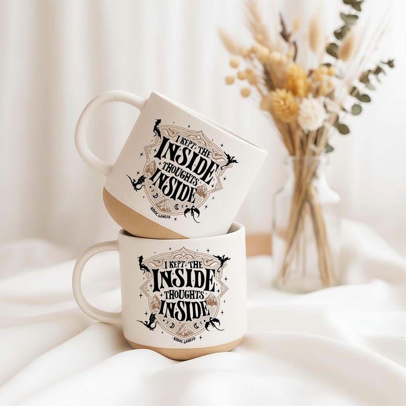 May include: Two off-white ceramic mugs stacked vertically, each featuring a black and gold graphic design with the text I KEPT THE INSIDE THOUGHTS INSIDE. The mugs have a smooth handle and a natural, unglazed tan base, showcasing a detailed illustration.