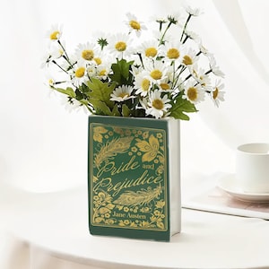 Può includere: Un vaso in ceramica bianca a forma di libro, con il titolo "Pride and Prejudice" in lettere dorate su sfondo verde. Contiene un bouquet di margherite bianche con centri gialli e foglie verdi. Sullo sfondo, una tazza bianca.