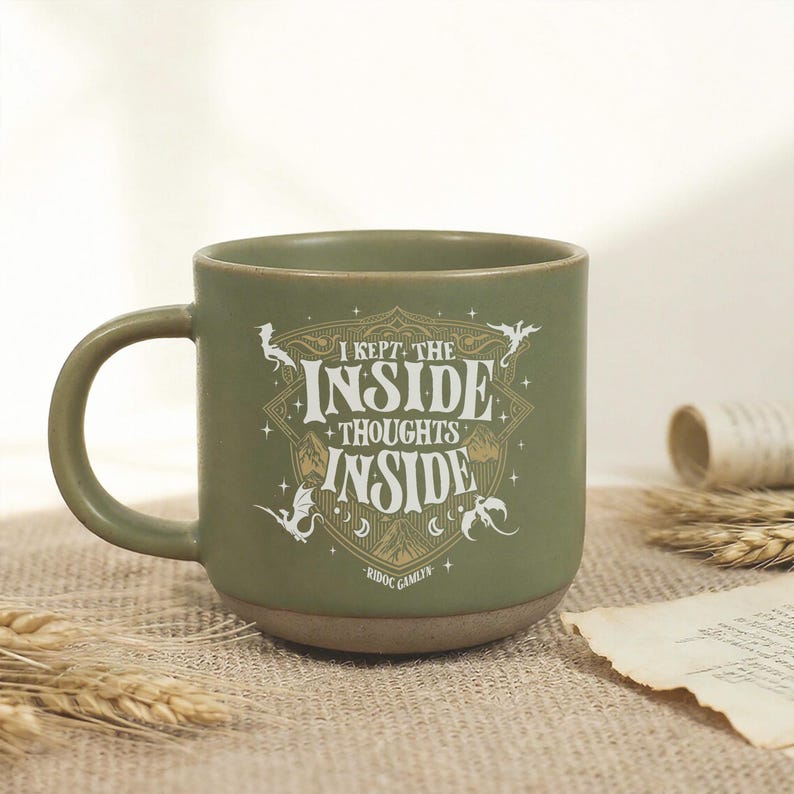 May include: A sage green ceramic mug featuring a white graphic design with the text I KEPT THE INSIDE THOUGHTS INSIDE and RIDOC GAMLYN. The design includes illustrations of dragons, stars, and crescent moons arranged within a shield-like emblem.