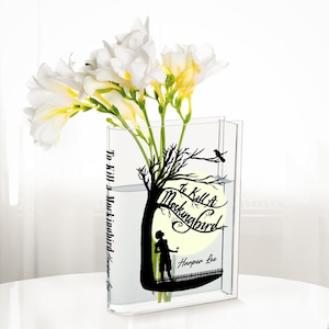 May include: A clear acrylic vase shaped like a book, featuring a black silhouette design from "To Kill a Mockingbird" by Harper Lee. The vase holds white and yellow freesia flowers, creating a decorative literary theme.