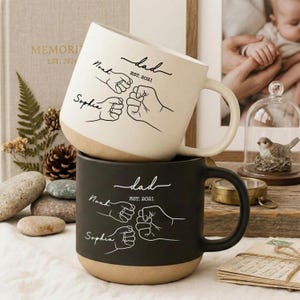 Peut inclure: Deux mugs en céramique, un blanc et un noir, empilés l'un sur l'autre. Le mug blanc a une base beige et une anse incurvée. Les deux mugs présentent le mot "dad", les noms "Noah" et "Sophia" et un motif de poing.