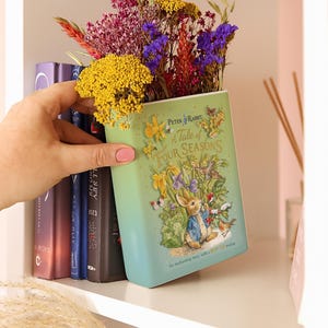 May include: A Peter Rabbit "A Tale of Four Seasons" book-shaped vase filled with colorful flowers. The vase is light green with illustrations of Peter Rabbit and other characters. The vase is on a white shelf with other books.
