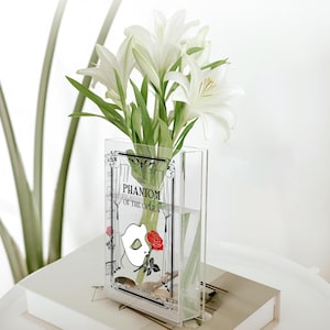May include: Clear acrylic vase featuring a graphic from "The Phantom of the Opera" and a bouquet of white lilies. The vase is rectangular and filled with water. The lilies have green stems and leaves.