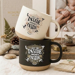 May include: Two ceramic mugs, one cream and one black, each featuring a shield-shaped graphic with the text I KEPT THE INSIDE THOUGHTS INSIDE and small dragon illustrations. The mugs have a smooth finish and a rounded base with a natural tan bottom.