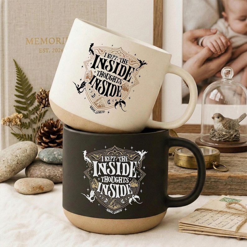 May include: Two ceramic mugs, one cream and one black, each featuring a shield-shaped graphic with the text I KEPT THE INSIDE THOUGHTS INSIDE and small dragon illustrations. The mugs have a smooth finish and a rounded base with a natural tan bottom.