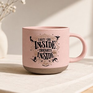 May include: A light pink ceramic mug with a speckled finish and a dark brown base. The front features black text that reads I KEPT THE INSIDE THOUGHTS INSIDE, surrounded by a decorative frame, small dragon silhouettes, stars, and the text RIDOC GAMLYN.
