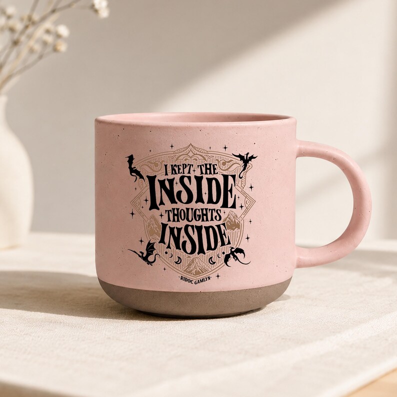 May include: A light pink ceramic mug with a speckled finish and a dark brown base. The front features black text that reads I KEPT THE INSIDE THOUGHTS INSIDE, surrounded by a decorative frame, small dragon silhouettes, stars, and the text RIDOC GAMLYN.
