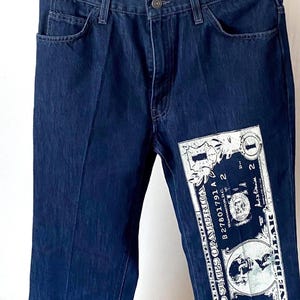 Op de afbeelding: Donkerblauwe denim jeans met een grote afbeelding van een dollarbiljet op de voorkant. De jeans hangen aan een zwarte hanger tegen een neutrale achtergrond.