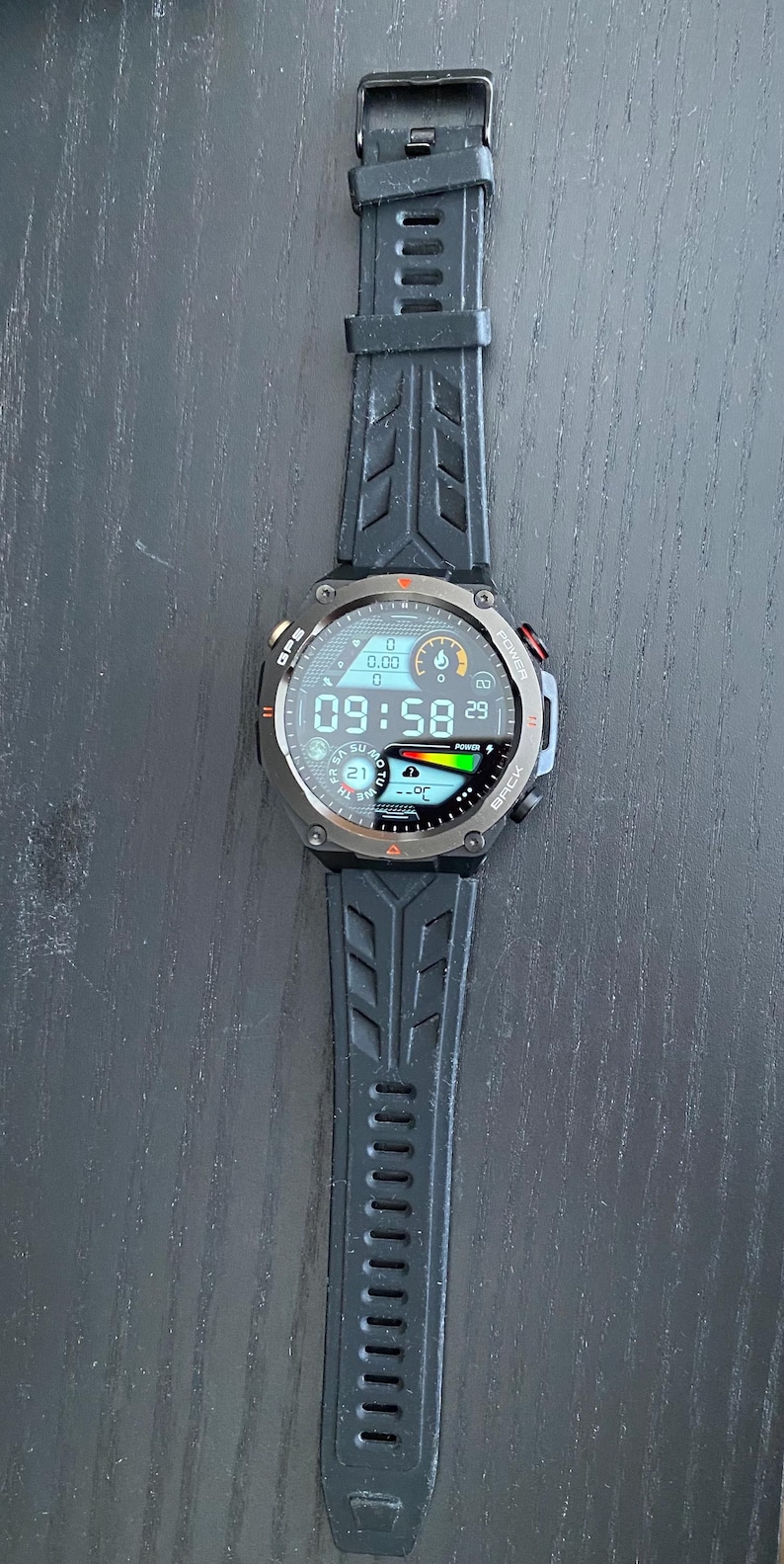 May include: Black smartwatch with a digital display showing the time as 09:58. The watch has a black strap with a textured design. The watch face displays various data points, including the date and battery life.