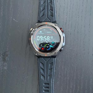 May include: Black smartwatch with a digital display showing the time, date, and weather. The watch has a black strap with a textured design. The watch face displays the time as 09:58. The watch has a metal bezel and buttons on the side.