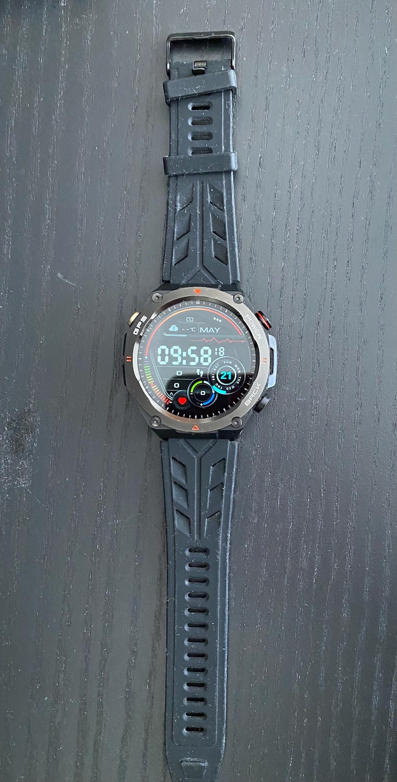 May include: Black smartwatch with a digital display showing the time, date, and weather. The watch has a black strap with a textured design. The watch face displays the time as 09:58. The watch has a metal bezel and buttons on the side.