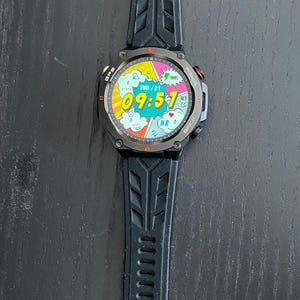 May include: Black smartwatch with a digital display showing the time 09:57 and the date. The watch has a black strap and a dark grey bezel. The watch face has a comic book style design.