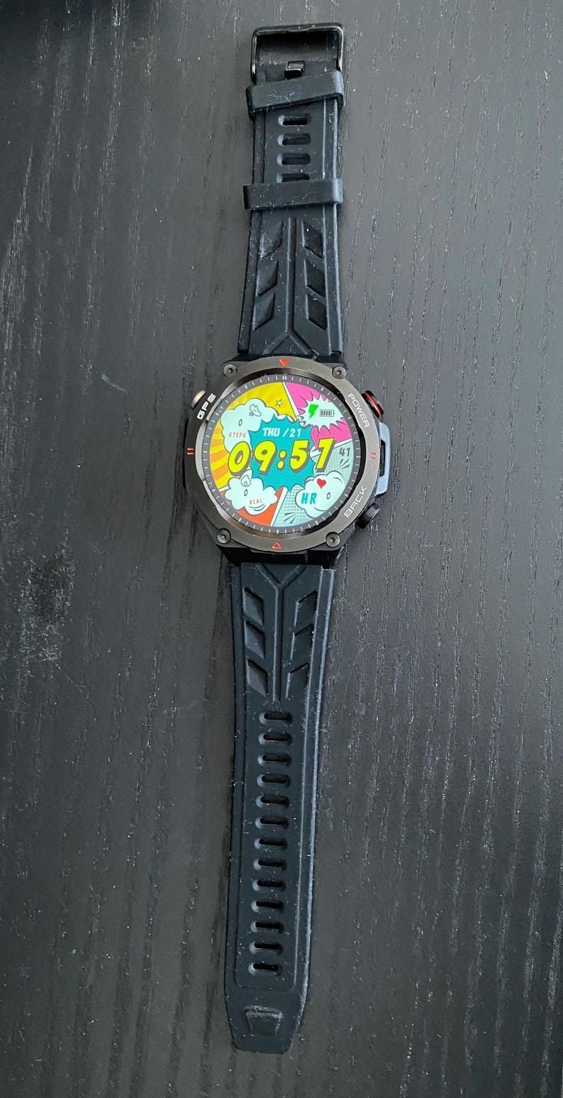May include: Black smartwatch with a digital display showing the time 09:57 and the date. The watch has a black strap and a dark grey bezel. The watch face has a comic book style design.