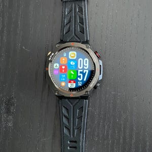 May include: Black smartwatch with a digital display showing the time as 09:57. The watch has a black strap with a textured design. The screen displays icons for various functions, including GPS and power.