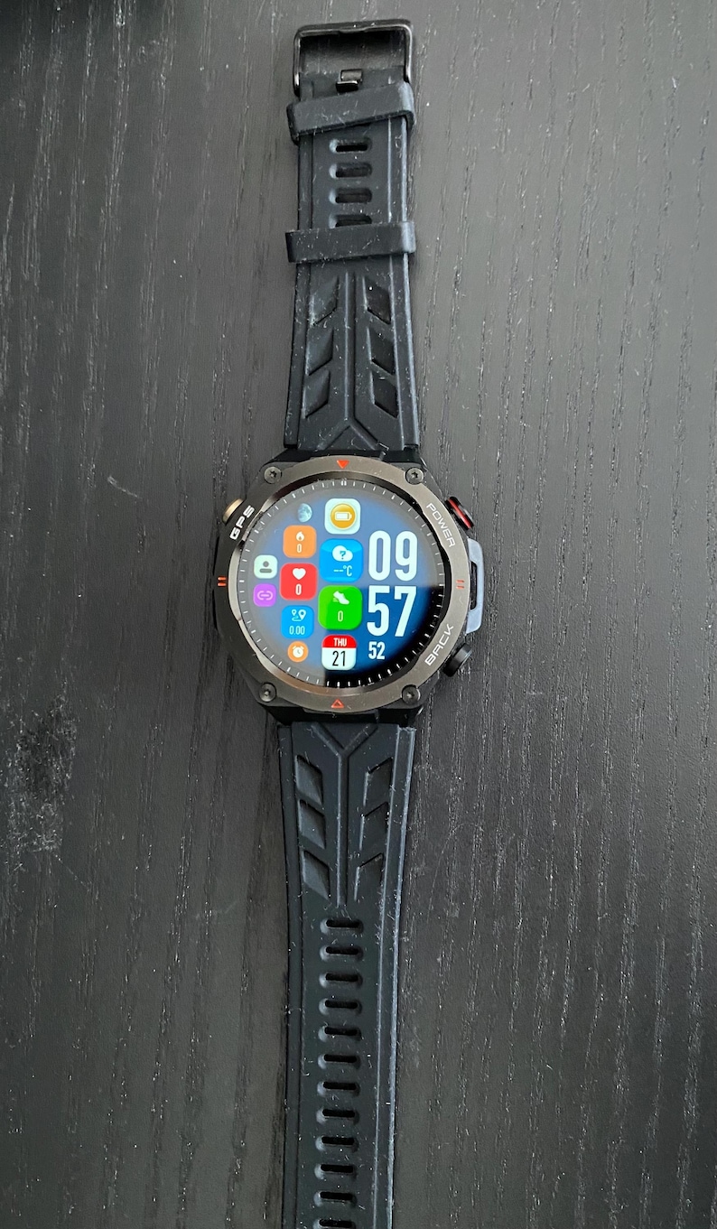 May include: Black smartwatch with a digital display showing the time as 09:57. The watch has a black strap with a textured design. The screen displays icons for various functions, including GPS and power.