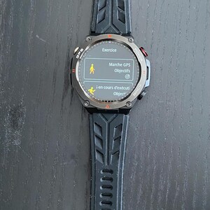 May include: A black smartwatch with a textured black strap. The watch face displays the word "Exercice" and other French text. The watch has a round face with red and silver accents. The strap is made of a flexible material.