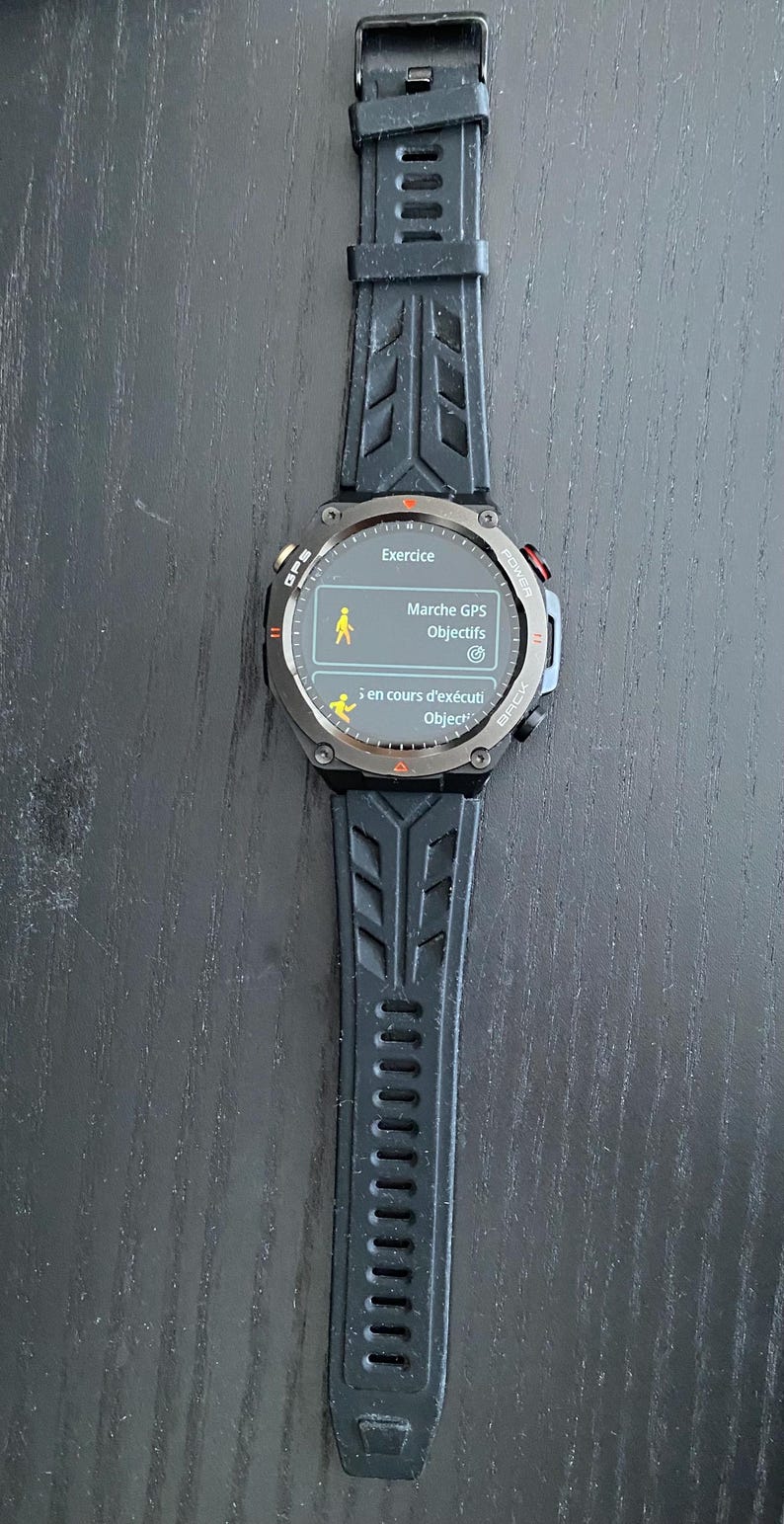 May include: A black smartwatch with a textured black strap. The watch face displays the word "Exercice" and other French text. The watch has a round face with red and silver accents. The strap is made of a flexible material.