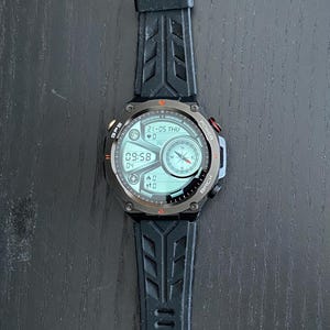 May include: Black smartwatch with a digital display showing the date, time, and other metrics. The watch has a black rubber strap with a textured design. The watch face has a compass and other data.