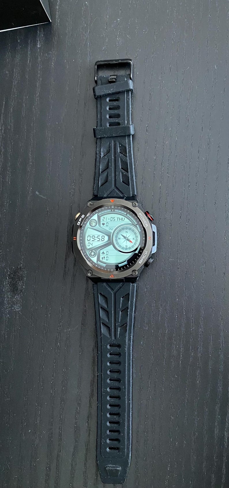 May include: Black smartwatch with a digital display showing the date, time, and other metrics. The watch has a black rubber strap with a textured design. The watch face has a compass and other data.