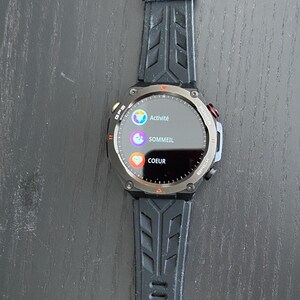 May include: Black smartwatch with a round face displaying "Activit&eacute;", "Sommeil", and "Coeur" in French. The watch has a black strap and a silver bezel. The watch is on a dark surface.