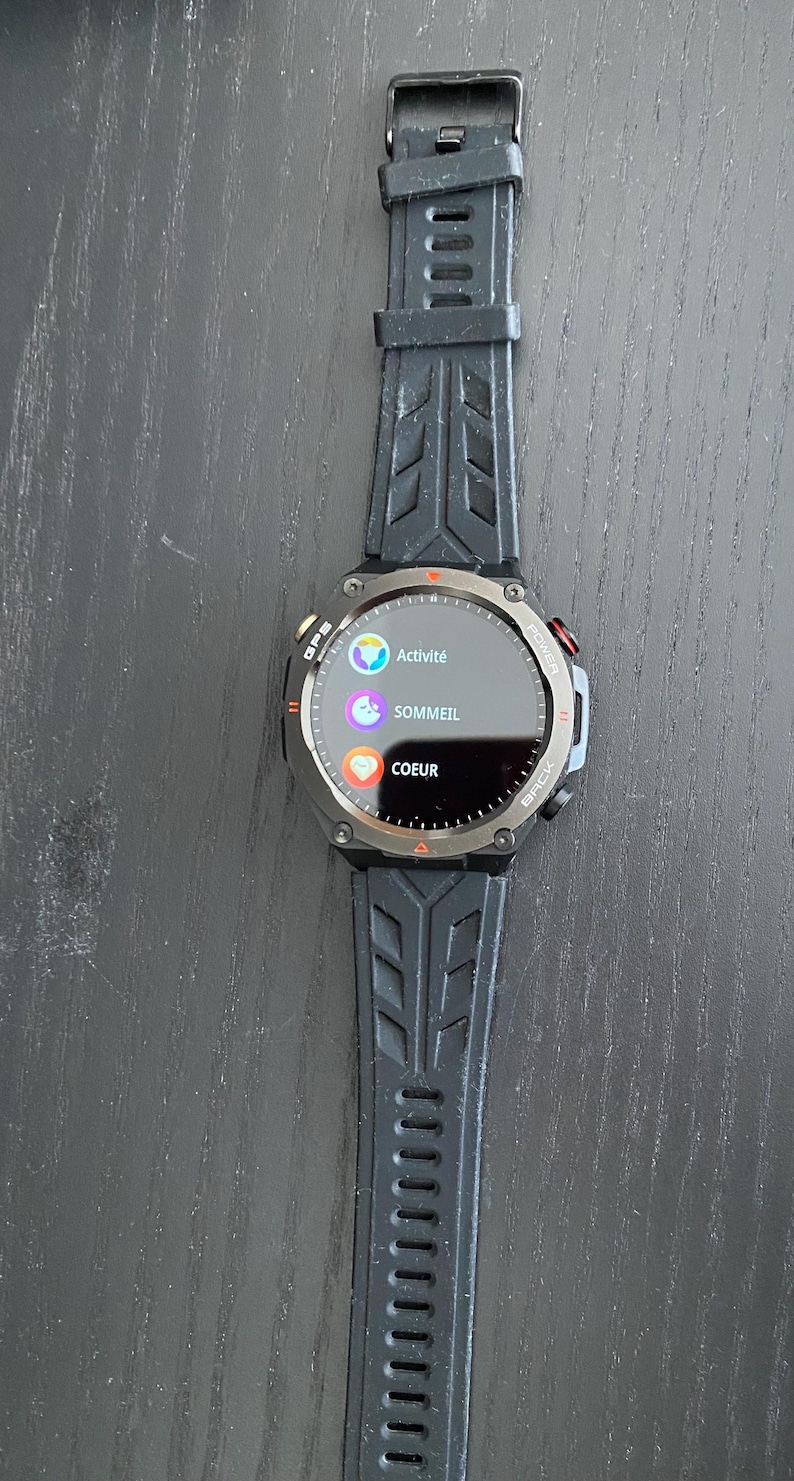 May include: Black smartwatch with a round face displaying "Activit&eacute;", "Sommeil", and "Coeur" in French. The watch has a black strap and a silver bezel. The watch is on a dark surface.