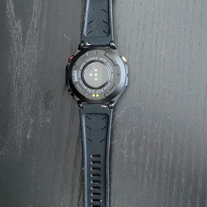 May include: Black smartwatch with a textured black strap. The watch face is black with a circular design and a few coloured accents. The watch is designed for outdoor and sport activities.
