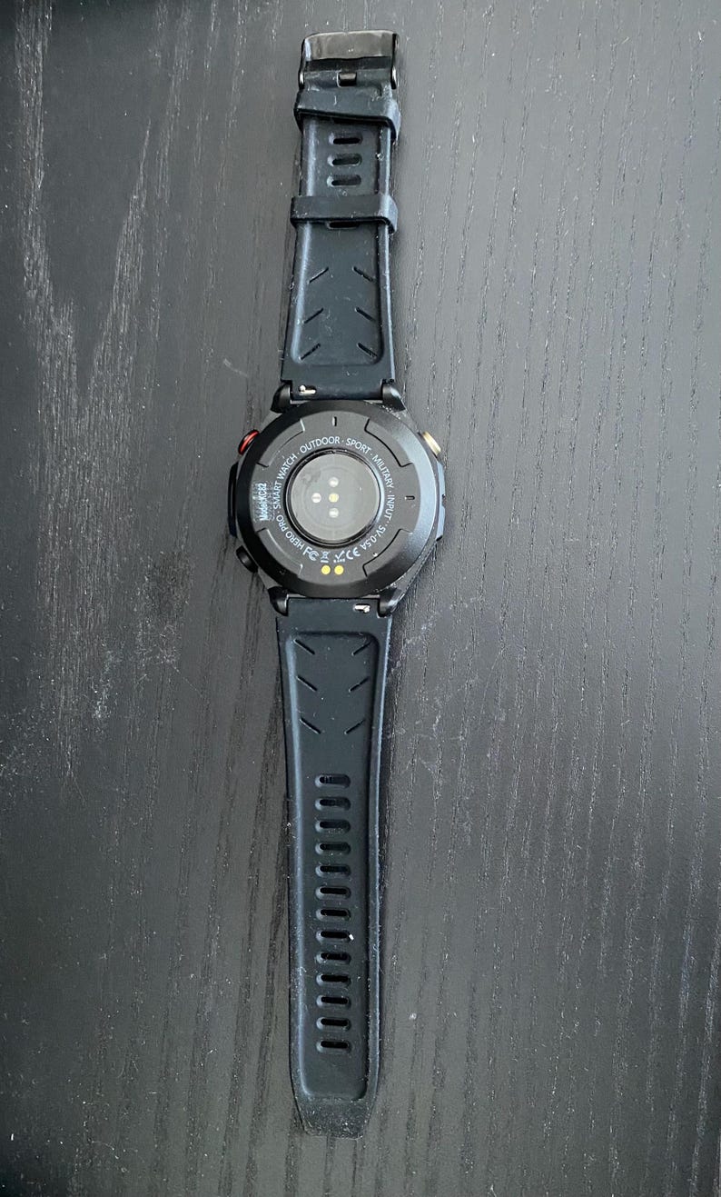 May include: Black smartwatch with a textured black strap. The watch face is black with a circular design and a few coloured accents. The watch is designed for outdoor and sport activities.