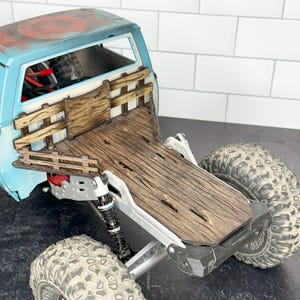 May include: A detailed view of a model truck bed with a weathered wood finish. The truck bed is light blue with a distressed paint effect. The tires are large and off-road style. The model has a realistic appearance.