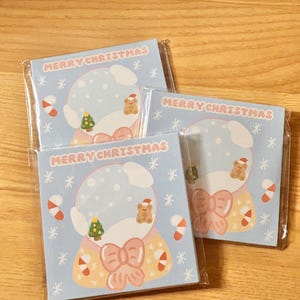 Könnte beinhalten: Drei quadratische Aufkleber zum Thema Weihnachten in Klarsichtverpackung. Jeder Aufkleber zeigt ein Schneekugel-Design mit einem Teddybären, einem Weihnachtsbaum und dem Schriftzug "MERRY CHRISTMAS" in Rosa. Zuckerstangen und Schneeflocken vervollständigen das festliche Design.