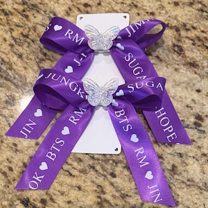 Può includere: Due fiocchi di raso viola con testo bianco e cuori. Ogni fiocco presenta un ornamento a farfalla trasparente. Il testo include nomi e iniziali. I fiocchi sono attaccati a una carta bianca.
