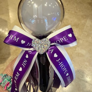 May include: A black BTS light stick featuring a clear spherical top with the BTS logo inside. A purple and white ribbon bow is tied around the handle, decorated with a central silver heart-shaped rhinestone and the names of the group members.