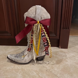 May include: A white western-style boot featuring intricate silver embroidery and glitter accents. The boot is adorned with a large maroon satin bow, gold ribbon, white lace trim, and a patterned ribbon featuring small circular logos and spear graphics.
