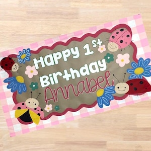 Op de afbeelding: Een rechthoekige banner met een roze en witte geruite achtergrond. De banner heeft de tekst "Happy 1st Birthday Annabel" in witte en roze letters. De banner is versierd met cartoon lieveheersbeestjes en bloemen.