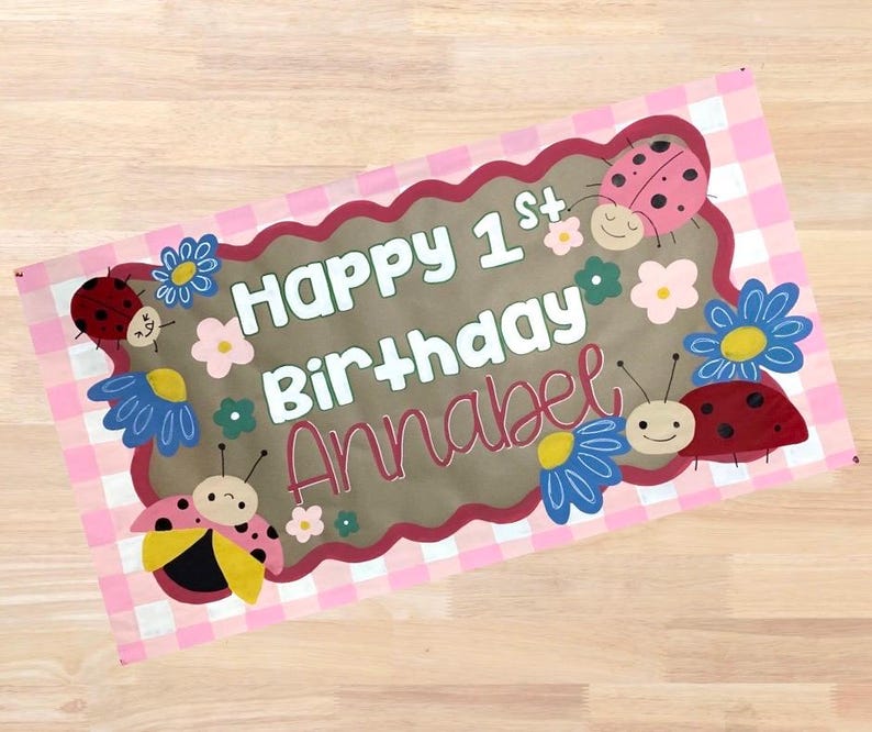 Op de afbeelding: Een rechthoekige banner met een roze en witte geruite achtergrond. De banner heeft de tekst "Happy 1st Birthday Annabel" in witte en roze letters. De banner is versierd met cartoon lieveheersbeestjes en bloemen.